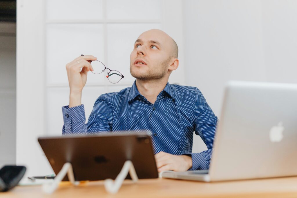 A fellow in an office holding his glasses and looking up thoughtfully.